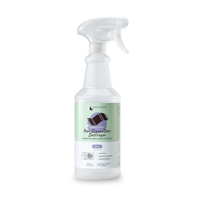 A transparent spray bottle, kin+kind Pet Pee + Stain + Odor Remover for Carpets and Fabrics, features a dog silhouette and paw print. With a green label and white trigger nozzle, it uses plant technology and natural ingredients to eliminate pet odors effectively.
