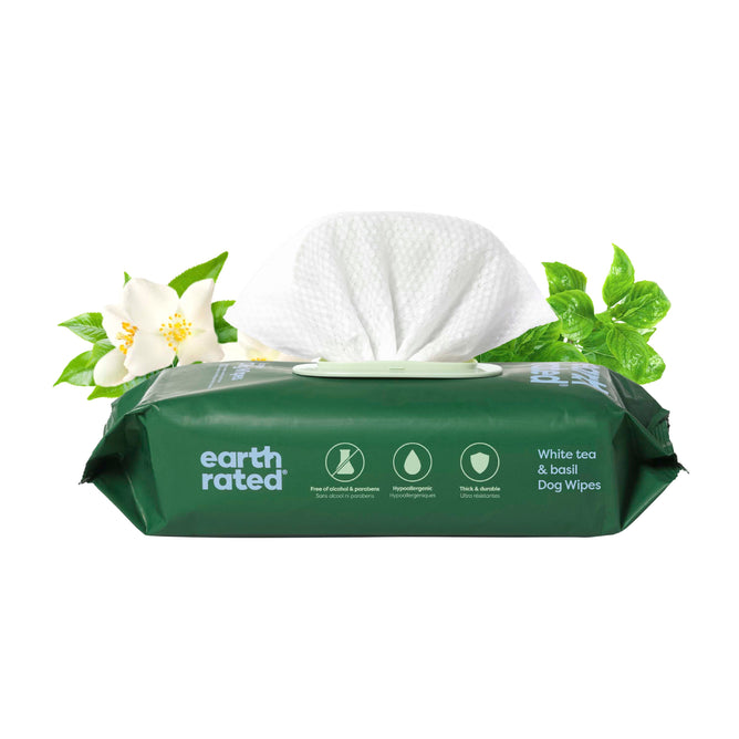 A green pack of Earth Rated Dog Grooming Wipes – Extra Thick, White Tea Basil Scented is shown with a wipe being pulled out. Jasmine flowers and basil leaves are behind the pack. Icons highlight hypoallergenic, plant-based, and thick wipes.