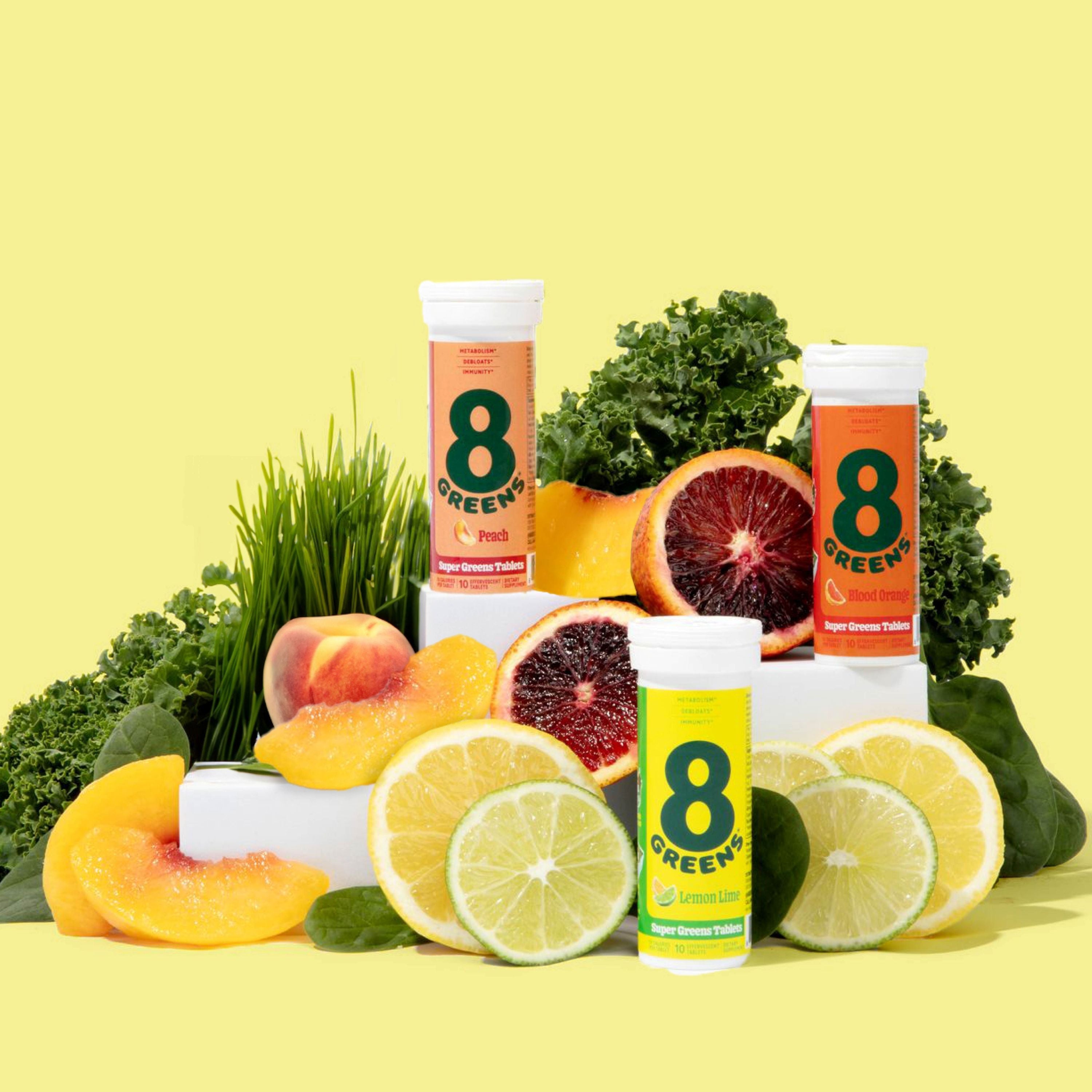 Three tubes of 8Greens Super Greens Tablet - Variety Pack in Peach, Blood Orange, and Lemon Lime flavors are surrounded by citrus slices, kale, spinach, and peach. These dietary supplements support metabolism and boost immunity against a light yellow backdrop.