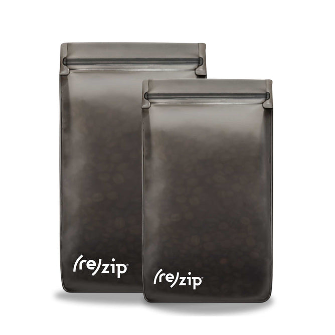The Coffee/Tea Storage Bag Kit includes two reusable, semi-transparent dark gray bags with the brand (re)zip in white at the bottom. They stand upright and have a zip-lock top, perfect for maintaining freshness of coffee beans or tea leaves.