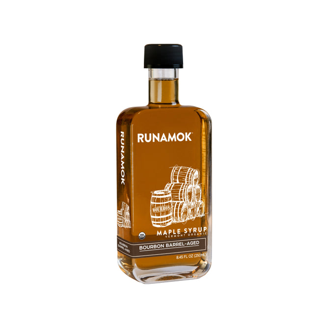 A clear glass bottle of Runamok Bourbon Barrel-Aged Maple Syrup, featuring barrel illustrations and a black cap, stands against a white background—crafted in bourbon barrels for rich flavor.