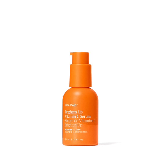 An upright orange bottle of Ursa Major Brighten Up Vitamin C Serum with a pump dispenser displays white and yellow text on a white background.