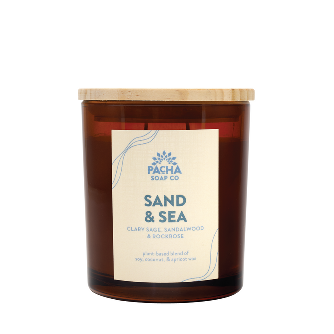 The Signature Candle - Sand & Sea by Pacha Soap Co. comes in a brown glass jar with a wooden lid, featuring a plant-based wax blend (soy, coconut, apricot) and an ocean-inspired scent of clary sage, sandalwood, and rockrose.