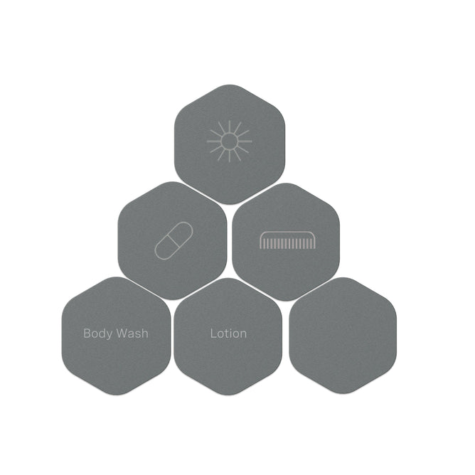 Six gray hexagonal Cadence capsules with interchangeable magnetic labels—Sun, Capsule, Comb, Body Wash, and Lotion—are arranged in a pyramid. Customize your travel system with the Cadence Labels - Set of 6.