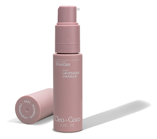 A pink bottle of Cleo & Coco Body + Hair Powder stands upright with its cap beside it. The label and cap display white text, highlighting this aluminum-free formula made from natural ingredients including arrowroot powder.