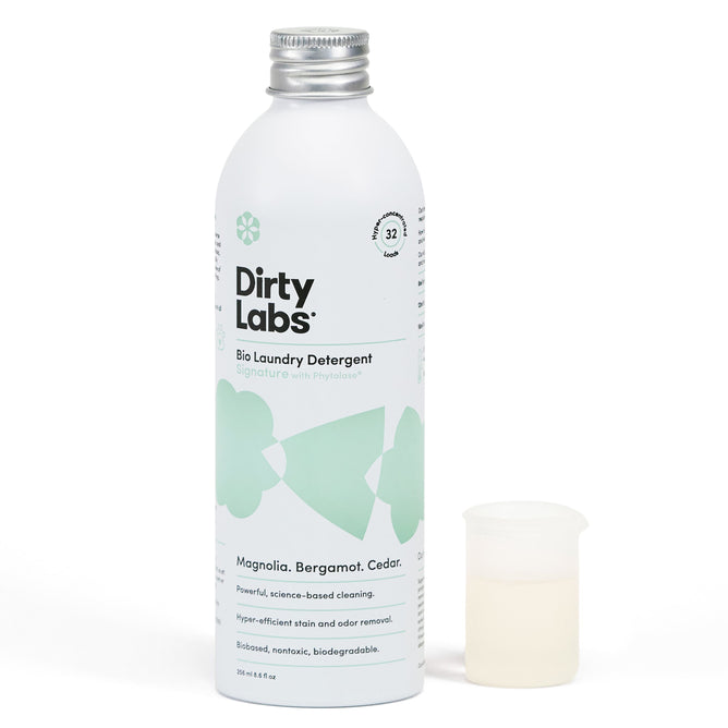 A bottle of Dirty Labs Bio Laundry Detergent with Phytolase® is shown next to a small measuring cup, featuring pale green graphics and promoting natural scents of magnolia, bergamot, and cedar.