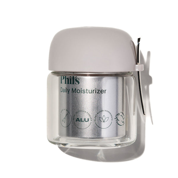 Phils Daily Moisturizer – First Phil comes in a clear glass jar with a light gray lid and metal clasp. The label notes aluminum packaging, plant-based ingredients, and nourishing marula oil plus hyaluronic acid.