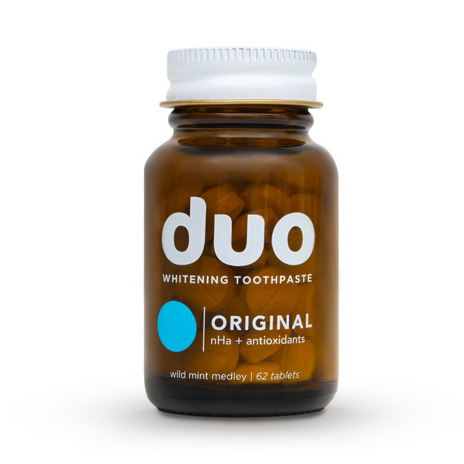 A brown glass bottle labeled duo Toothpaste Whitening Toothpaste Tablets contains 62 tablets featuring an Original nHa and antioxidants formula for oral health. The metal-capped bottle has a blue circle design, offering a wild mint medley with nano-hydroxyapatite benefits.