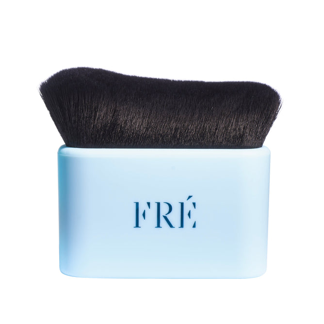The FRÉ Skincare Bronzing Body Brush features dense black bristles and a light blue rectangular handle with FRÉ on the front. Achieve a streak-free glow with this flat-topped must-have tool. The background is white.