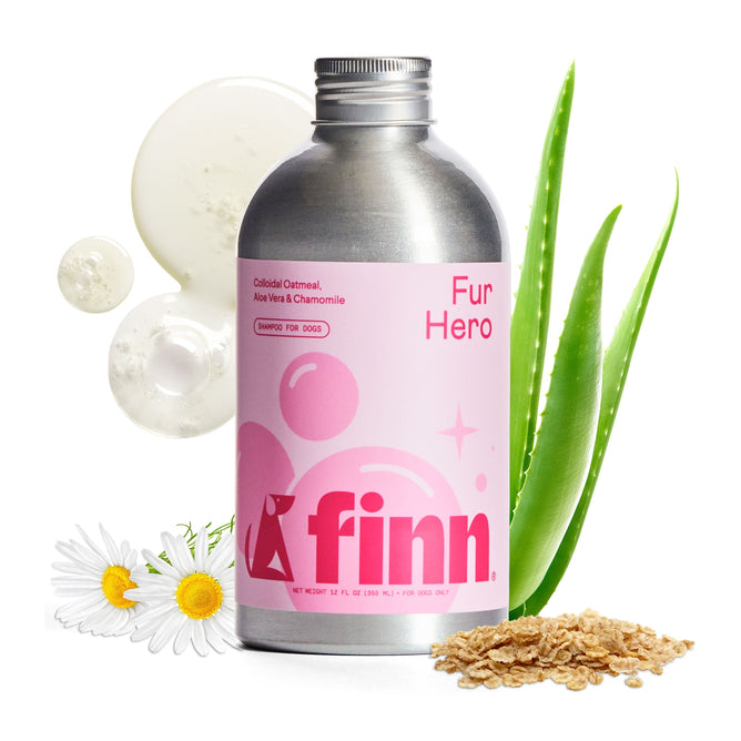 A silver and pink Finn Fur Hero Shampoo for Dogs bottle sits with chamomile, aloe vera, oatmeal, and creamy drops. This all-natural shampoo soothes sensitive canine skin using colloidal oatmeal, aloe vera, and chamomile.