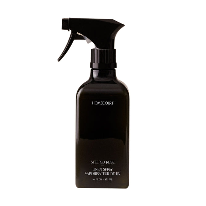 A black Homecourt Linen Spray bottle with white text stands upright against a plain white background, offering an eco-friendly fabric fragrance for your home.