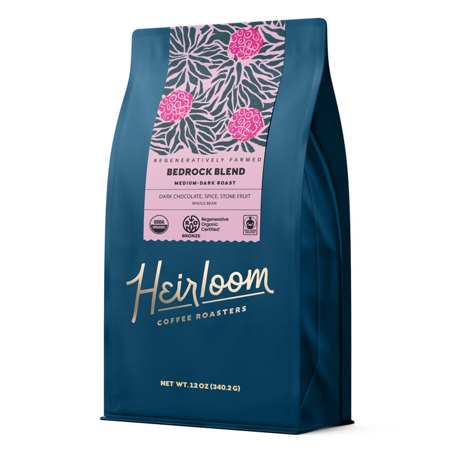 A blue bag of Heirloom Coffee Roasters Bedrock Blend ROC Whole Bean Coffee, a medium-dark roast with notes of bittersweet chocolate, spice, and stone fruit. Features pink label designs. Sourced from regenerative farms. Net wt: 12 oz (340.2 g).