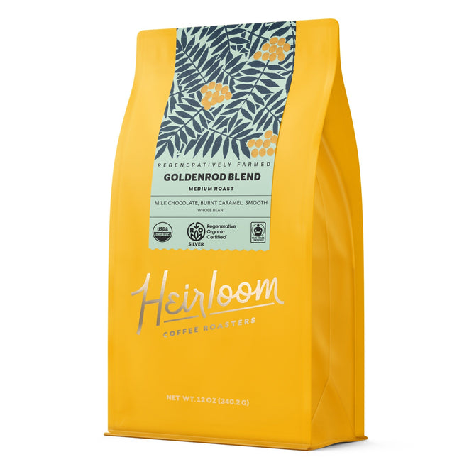 A yellow bag of Heirloom Coffee Roasters Goldenrod Blend ROC Whole Bean Coffee features a leafy floral design, Medium Roast label, Regenerative Organic Certified farming, and tasting notes of milk chocolate and burnt caramel.