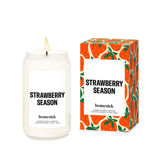 A lit Homesick Strawberry Season candle made of soy wax and natural scents sits in a glass jar next to its strawberry-patterned box. Enjoy this cruelty-free indulgence with matching branding.