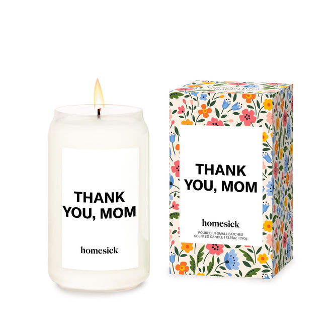 A lit Homesick Thank You, Mom Candle made from natural soy wax sits beside a matching floral box, both displaying the same heartfelt message.