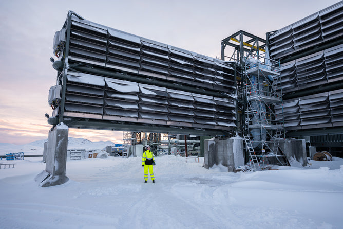 Wearing a bright yellow safety suit, an individual stands amid large metal structures by Grove Co., designed for Climate Impact through Direct Air Capture in a snowy industrial area. Snowy mountains loom under cloudy skies, emphasizing the urgency of combating climate change.