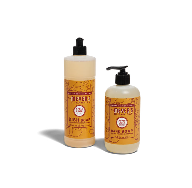 Two bottles from Mrs. Meyers Crisp & Clean Sinkside Set - Apple Cider: one dish soap with a flip cap and one hand soap with a pump, both with orange labels and floral designs, are shown standing on a white background.