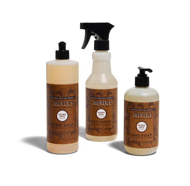 The Mrs. Meyers Happy Harvest Essentials Set in Acorn Spice includes dish soap, multi-surface spray, and hand soap, each with brown labels featuring acorn designs, displayed against a white background.
