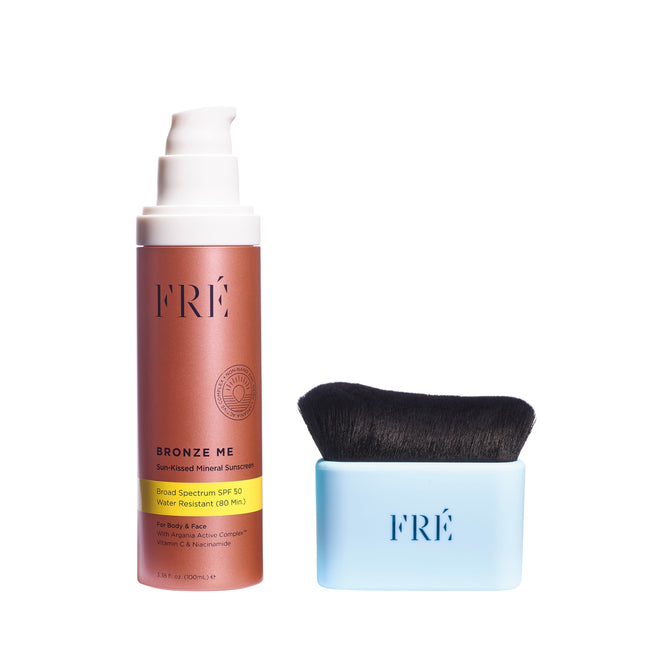 A bottle of FRÉ Skincare BRONZE ME Sun-Kissed Mineral Sunscreen SPF 50 with a white pump sits beside a blue applicator brush with soft black bristles, both on a white background. Includes free gift.