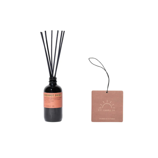 A bottle labeled Bergamot Shiso from the Alchemy Line Reed Diffuser by P.F. Candle Co., with black reeds and a brown tag featuring a sun design, sits on the left against a white background. It delivers mood-uplifting, nature-inspired scents.