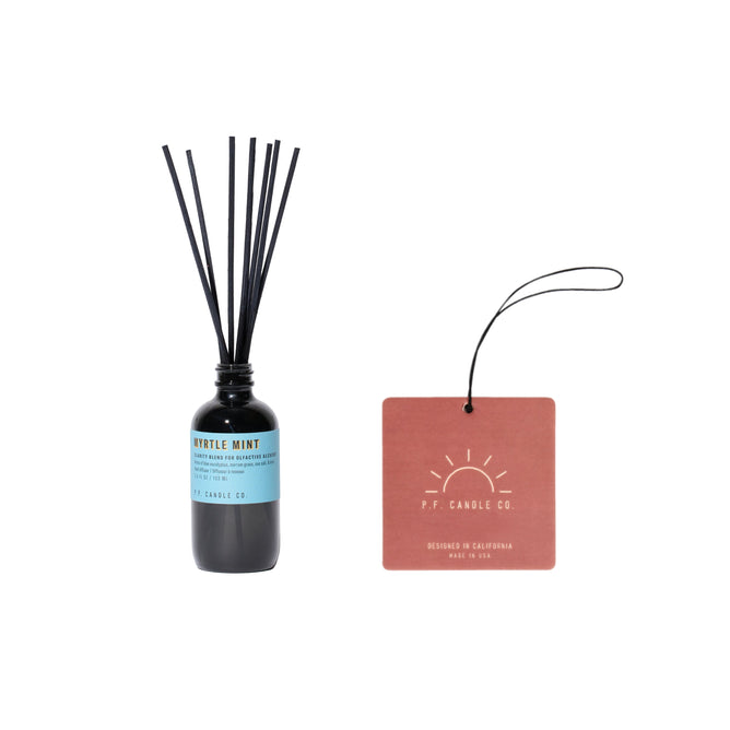 A dark glass reed diffuser bottle named Alchemy Line Reed Diffuser, Myrtle Mint - free gift stands elegantly with black reeds and a rustic orange tag reading P.F. Candle Co. Designed in California, this nature-inspired fragrance is made in the USA.