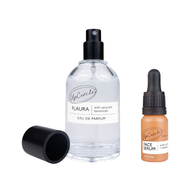 A bottle of UpCircles Upcycled Eau de Parfum labeled Flaura with a black cap detached is next to a small Face Serum dropper bottle, both infused with Orange Essential Oil and showcasing eco-conscious design.