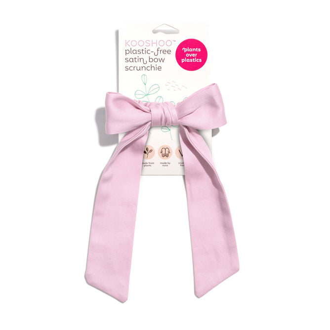 The KOOSHOO Satin Bow Scrunchie, made from plant-based satin and organic cotton, is shown on its packaging with a pink circle that reads “plants over plastics,” emphasizing its zero-plastic design.