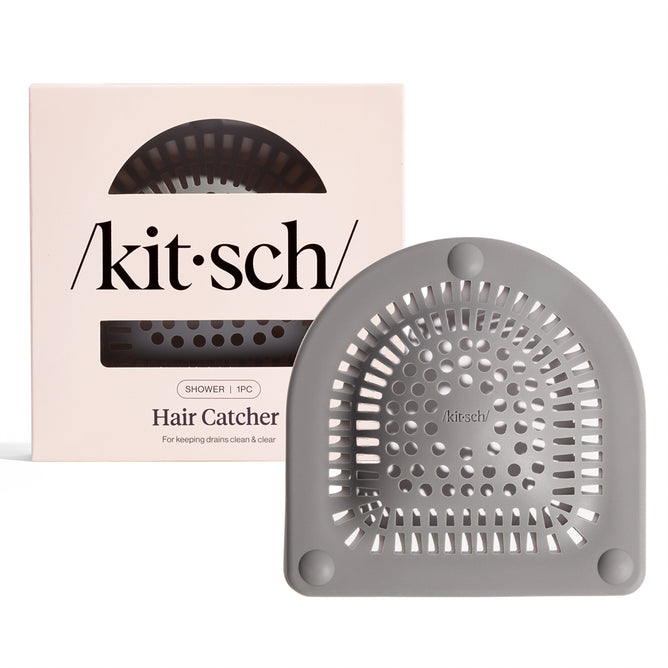 A Kitsch Hair Catcher ensures a clean shower by efficiently collecting hair. This gray, semi-circular device with a grid pattern is packaged in a cream-colored box labeled with the brand name, featuring a clear window to display the product.