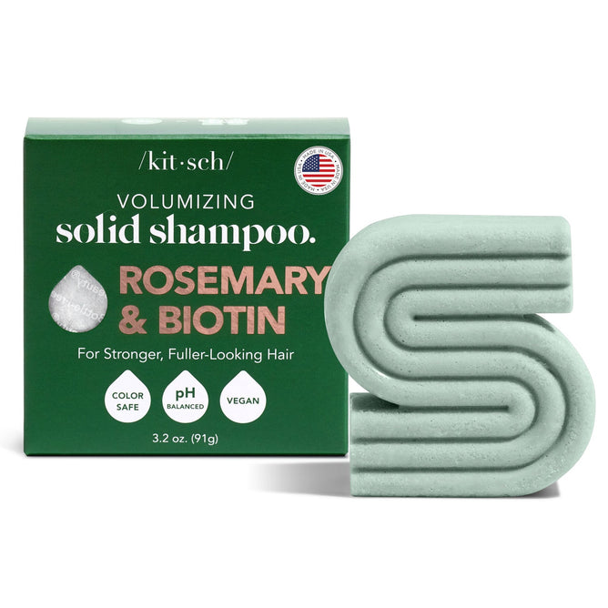 A green, S-shaped solid shampoo bar sits next to a box labeled Kitsch Rosemary & Biotin Solid Shampoo. This color-safe, pH-balanced vegan formula promotes hair growth and fuller-looking locks with biotin. A small USA flag symbol accompanies the text.