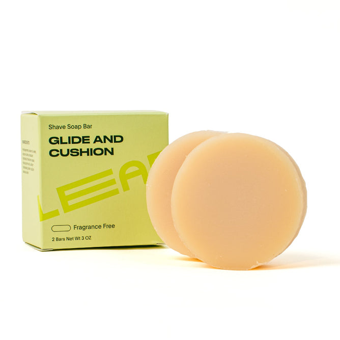 A box labeled Leaf Shave Shave Soap sits beside two round, beige bars. The natural soap creates a creamy lather and comes in a light green box marked Fragrance Free with 2 Bars Net Wt 3 OZ on the front.