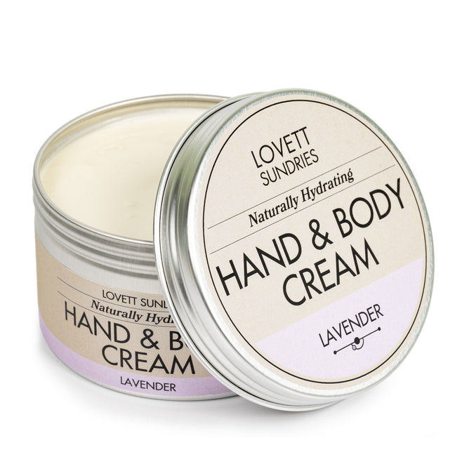A metal tin of Lovett Sundries Hand & Body Cream is open, showing the smooth cream inside, labeled with its soothing lavender scent. It features a minimalist design in calming beige and purple tones.