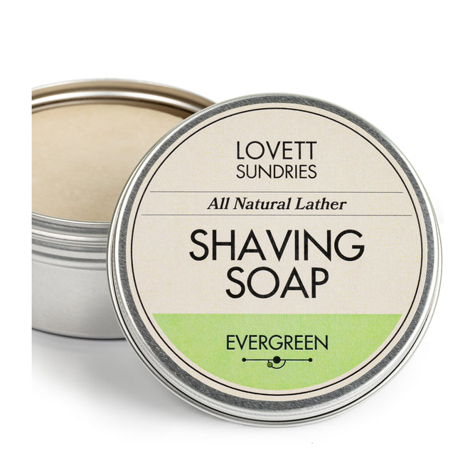A tin of Lovett Sundries Shaving Soap showcases a lid with All Natural Lather and Evergreen, emphasizing its natural ingredients. The open tin reveals a smooth, light-colored soap enriched with bentonite clay.