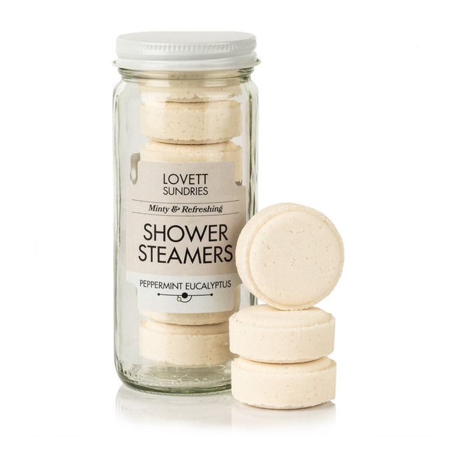 A jar of Lovett Sundries Shower Steamers contains round, beige tablets for aromatherapy with a soothing peppermint eucalyptus scent. Three extra tablets are stacked nearby.