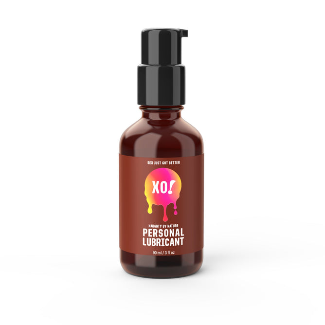 A 90 ml brown bottle of XO! Naughty by Nature Organic Lube from Here We Flo features a black pump dispenser and a pink and yellow label with bold white XO! and Personal Lubricant letters, offering a natural pH balance.