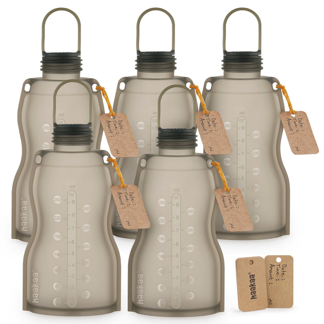 Six translucent gray Haakaa Reusable Breast Milk Storage Bags made from 100% food-grade, freezer-safe silicone, with measurement markings, black screw-top lids, carrying handles, and brown tags. Two extra tags are in the foreground.