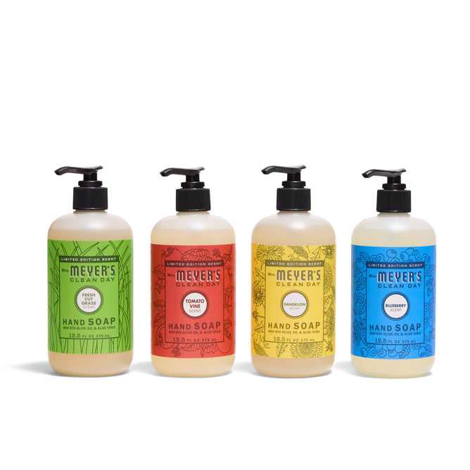 Four Mrs. Meyers Summer Hand Soap Collection bottles are lined up with pump tops and colorful labels for Basil (green), Tomato Vine (red), Patchouli (yellow), and Bluebell scents, showcasing the NEW vibrant lineup.