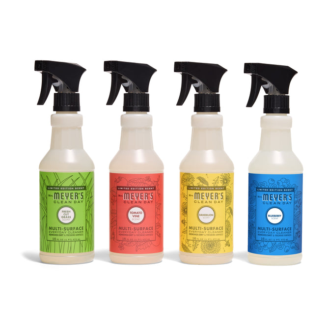 Four Mrs. Meyer’s Summer Multi-Surface Cleaner Collection spray bottles are lined up, each with a black spray top and colorful labels: green (parsley), red (tomato vine), yellow (marigold), and blue (bluebell) on white backgrounds.