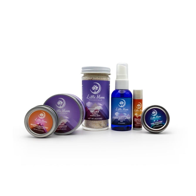 The Moms Essentials Survival Kit Gift Set by Little Moon Essentials is perfect for supermoms, featuring bath salts, a blue stress relief spray, lip balm, and various labeled tins—all elegantly displayed against a white background.