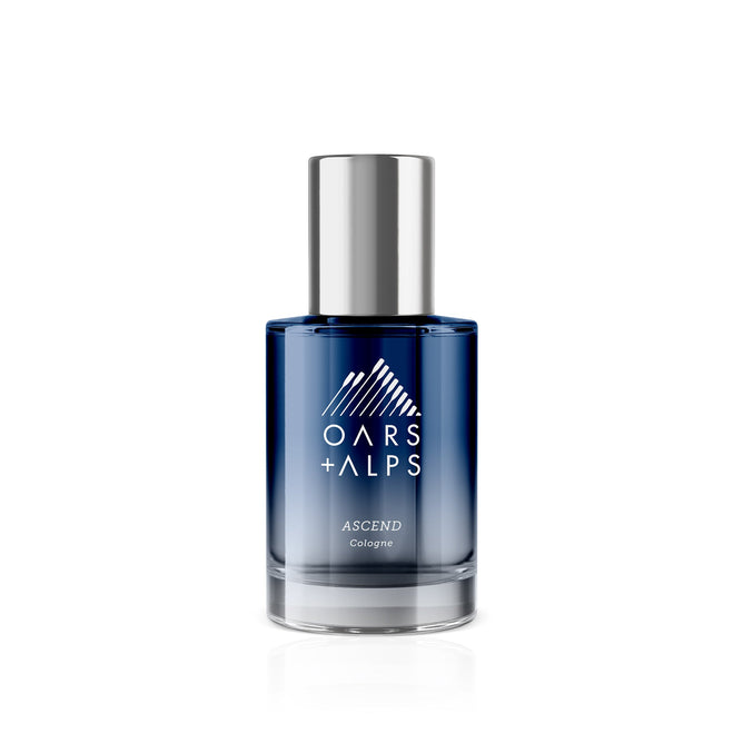 A blue glass bottle of Oars + Alps ASCEND Cologne with a silver cap features a white mountain logo and text on the front, set against a white background—a captivating cedar-scented fragrance by Oars + Alps.