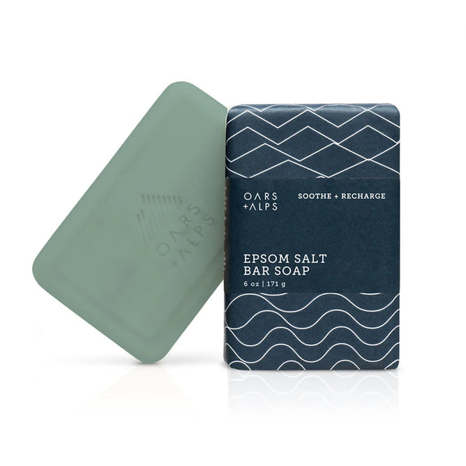 A green Oars + Alps Athlete Bar Soap with Epsom Salt sits beside its dark blue box, decorated with abstract white waves and Soothe + Recharge text, featuring a soothing eucalyptus spearmint scent.