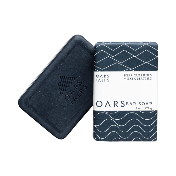 Two Oars + Alps Exfoliating Bar Soaps: one unwrapped, dark blue with an embossed logo; the other wrapped in blue paper with white geometric patterns and labeled Deep Cleaning + Exfoliating with volcanic sand.
