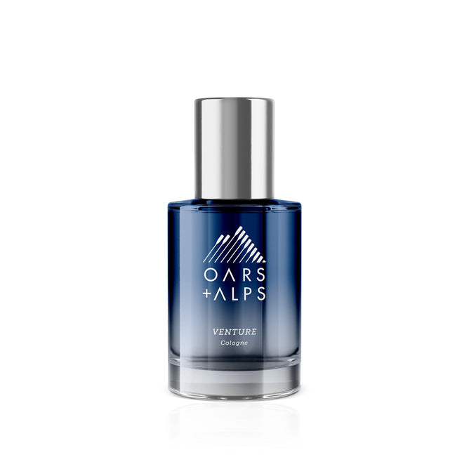 A blue glass bottle of Oars + Alps VENTURE Cologne with a silver cap and white logo is shown on a white background, reflecting the bergamot-inspired fresh marine and citrus fragrance of this signature scent.