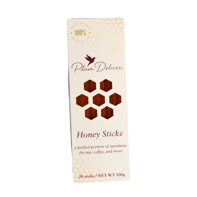 Plum Deluxe Honey Sticks come in a cream-colored box with gold accents, honeycomb patterns, and a bird logo. The packaging features honeycomb cutouts that reveal the tasty sticks inside—perfect for pairing with tea or wildflowers.