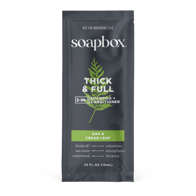 A 0.33 fl oz (10mL) sachet of Soapbox Thick & Full 2-in-1 - Sample Shampoo & Conditioner with biotin comes in dark gray packaging and a green Oak & Cedar Leaf label, promoting volumizing and strengthening benefits for fuller-looking hair.