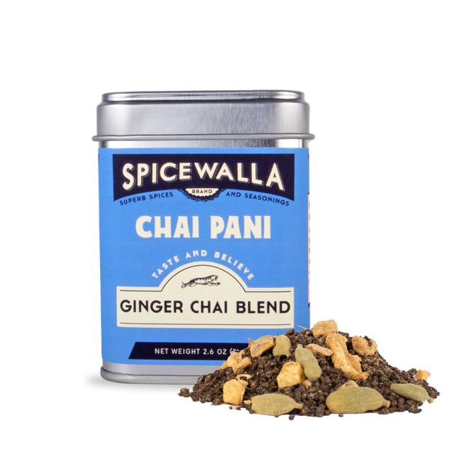 A tin of Spicewalla Chai Pani Ginger Chai with a blue label and silver lid sits beside loose black tea leaves dotted with ginger and cardamom.