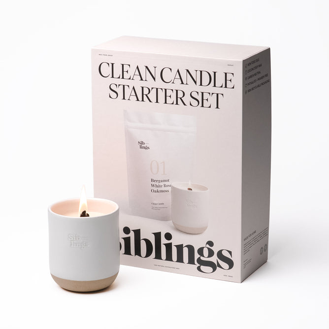 The Clean Candle Starter Set by Siblings includes a lit white candle in a minimalist Forever Vessel holder, beside a box labeled with the product name and featuring text and images of Siblings candles that describe their clean-burning properties and elevate your space with luxurious fragrance.