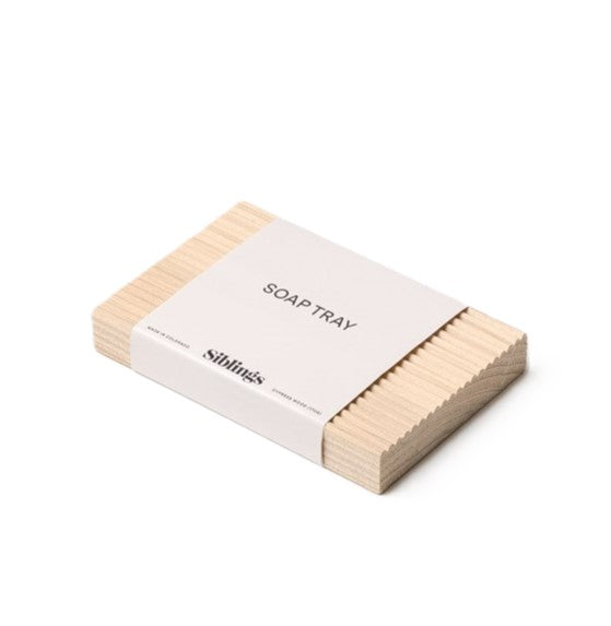 The Wooden Soap Tray by Siblings is rectangular, made from sustainably grown cypress with ridges for moisture resistance. Wrapped in a white band labeled WOODEN SOAP TRAY and Siblings, it has a light minimalistic design.