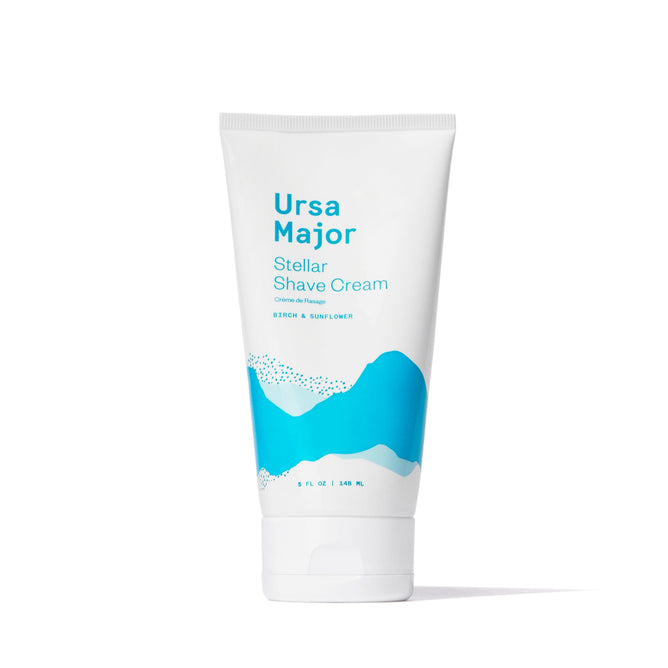 A white tube of Ursa Major Stellar Shave Cream featuring blue abstract mountain graphics stands upright on a plain white background. The label notes its “Cedar & Sunflower” botanical scent and size: 5 FL OZ / 148 mL.