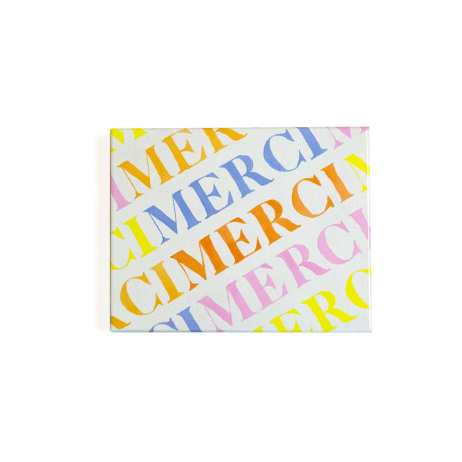 The Supra Endura Seed Card features MERCI repeated diagonally in pastel colors on a white canvas. Made from seed-embedded paper, it adds a unique touch—plant it to watch your gratitude grow.