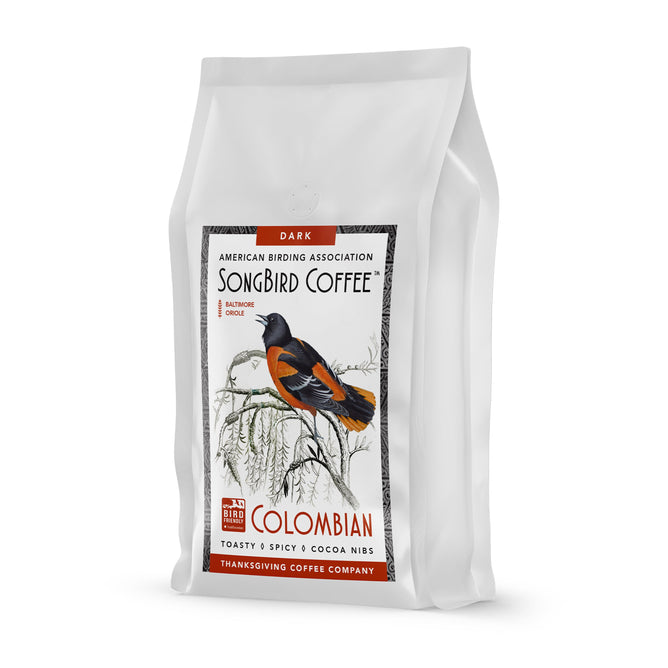 The white Songbird Colombian Dark bag by Thanksgiving Coffee features a Baltimore oriole illustration, touting its dark Colombian roast with notes of toast, spice, and cocoa nibs while supporting Bird Friendly Coffee via the American Birding Association.